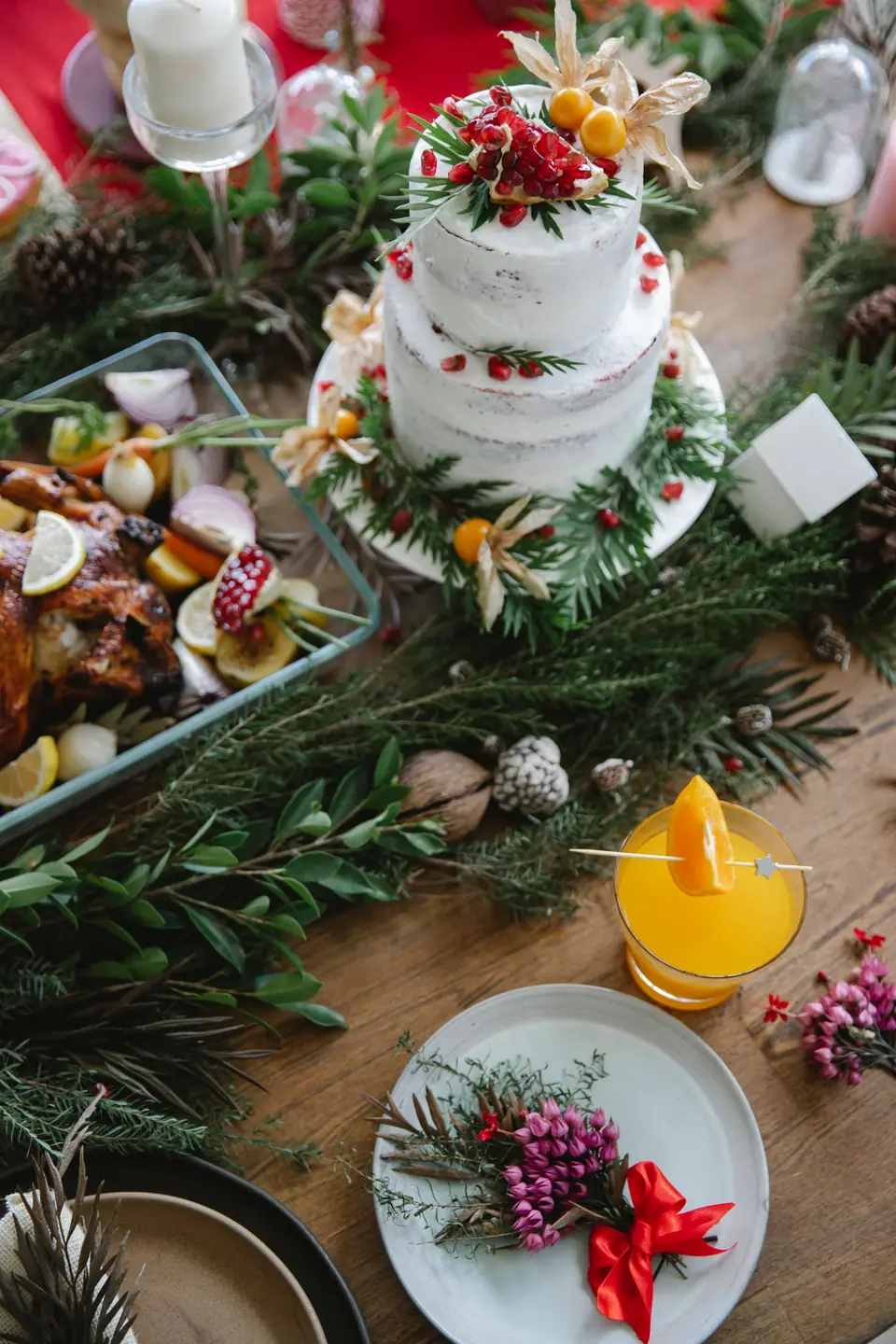 A tiered white cake with pomegranate and greenery on a Christmas table beside a roast and a drink