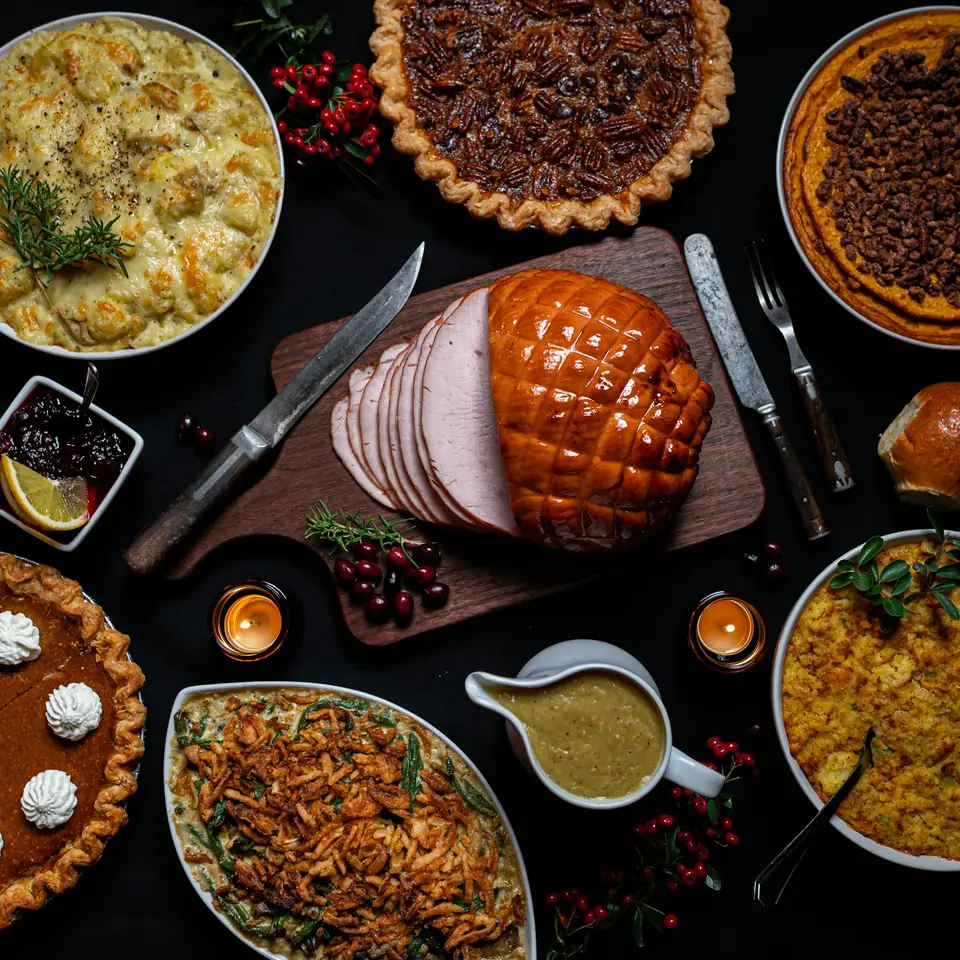 A glazed ham on a dark table surrounded by pies, gravy, and side dishes