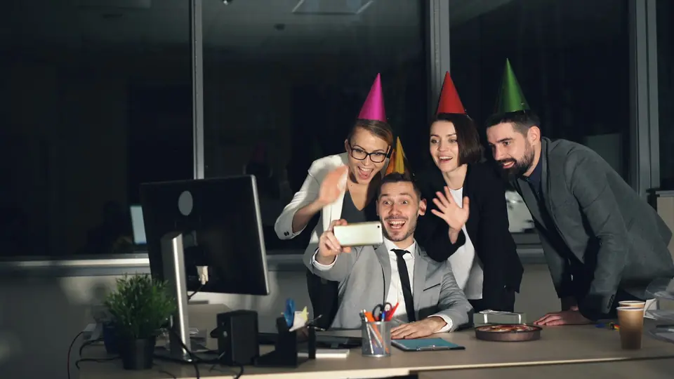 Coworkers in party hats crowding around a desk for a selfie